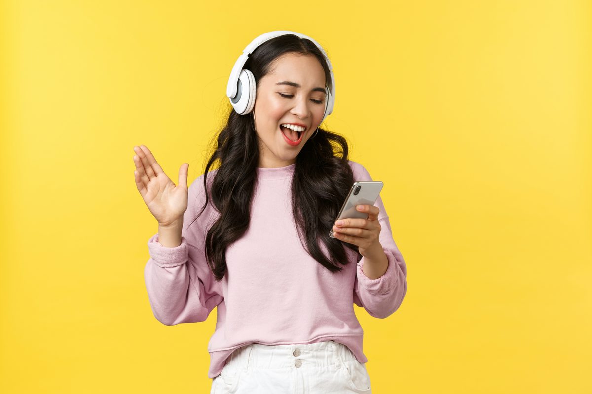 A woman in a pink sweater on a bright yellow background, dancing and singing along to music with white headphones